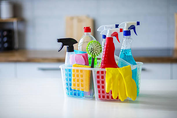 A plastic basket filled with various cleaning supplies including spray bottles, sponges, rubber gloves, and brushes placed on a white kitchen countertop. The background shows a modern kitchen with wooden and white cabinets, perfect for illustrating home cleaning, hygiene, or housekeeping concepts.